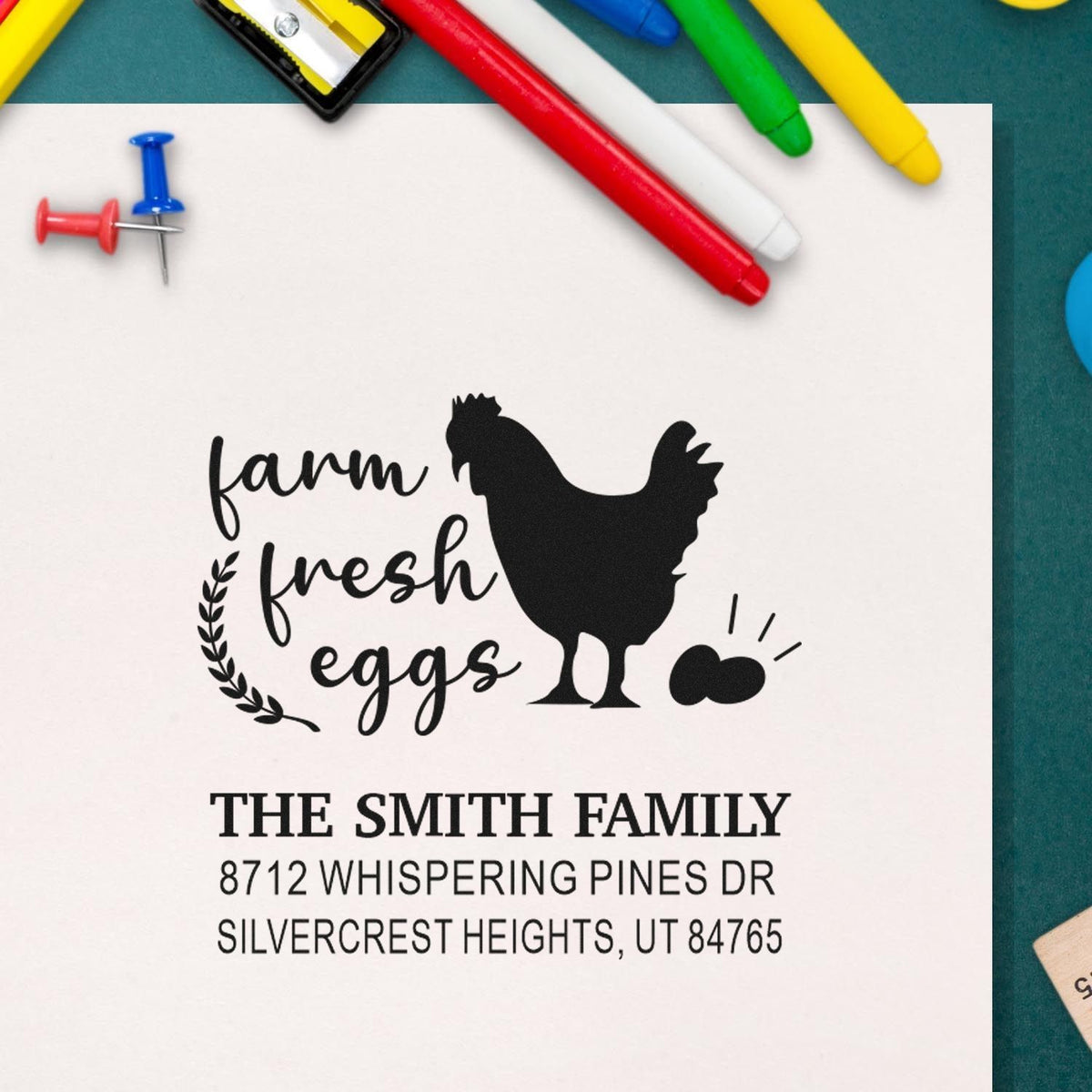 Custom farm fresh eggs return address stamp with chicken silhouette, The Smith Family address, displayed with colorful pens on a desk.