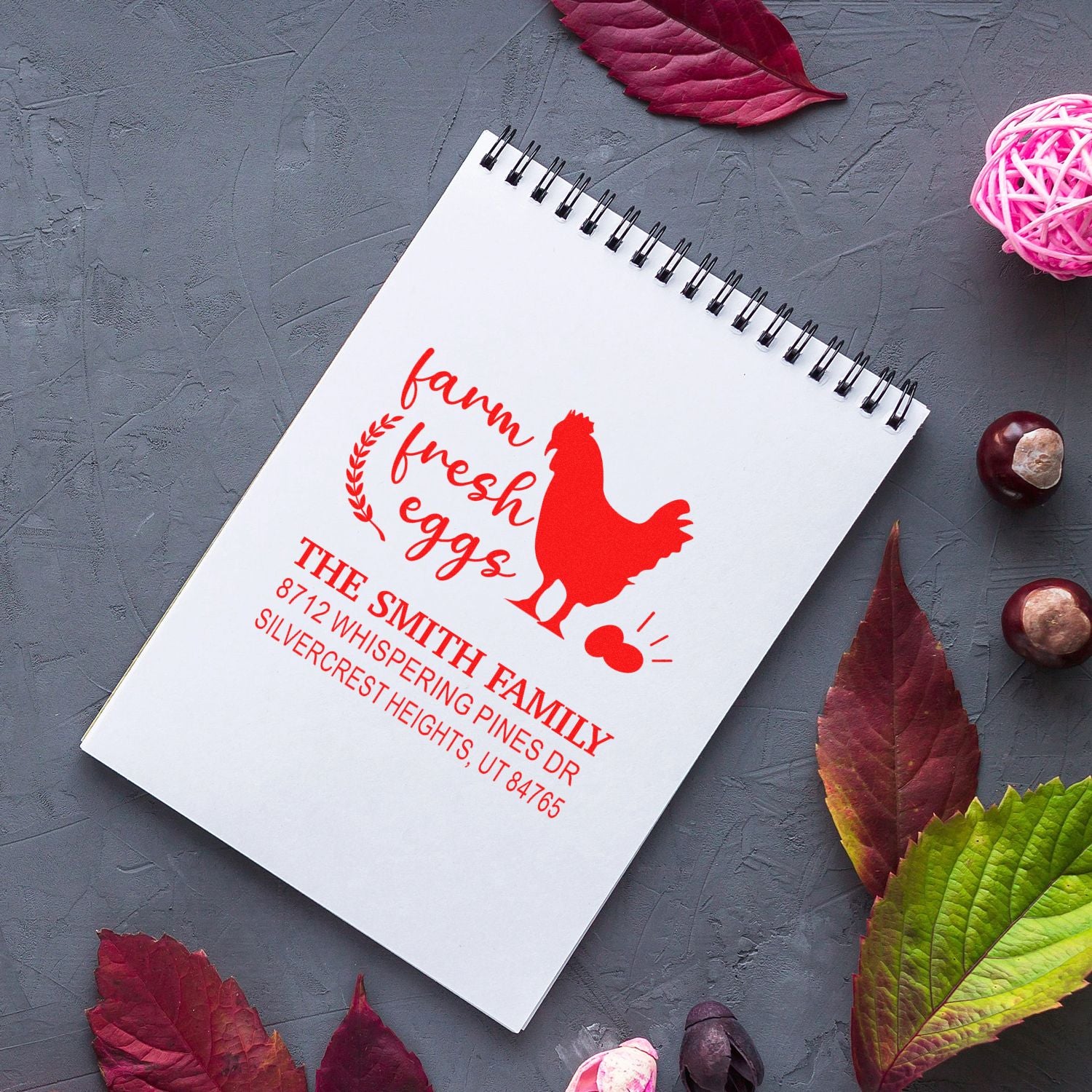 Custom red farm fresh eggs address stamp on spiral notebook, with rooster design, on gray tabletop surrounded by fall leaves and decor.