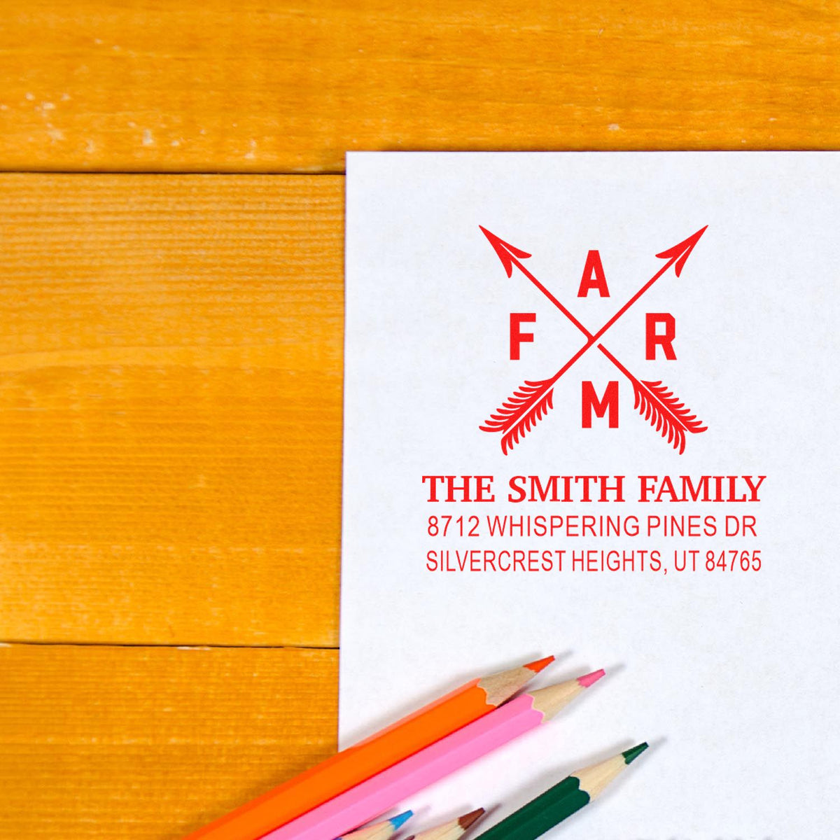 Custom family address stamp on white paper with red monogram, set on a wooden desk beside colored pencils.