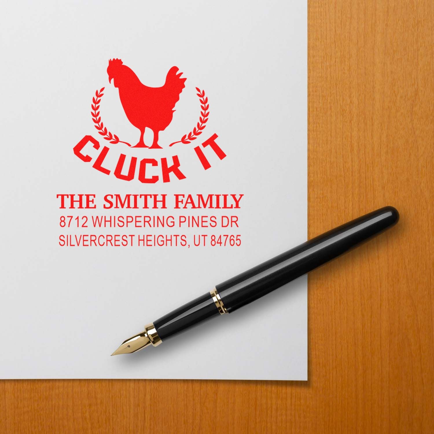 Custom red Cluck It family address stamp with rooster logo, The Smith Family text, beside a black fountain pen on a white sheet over a wood desk.
