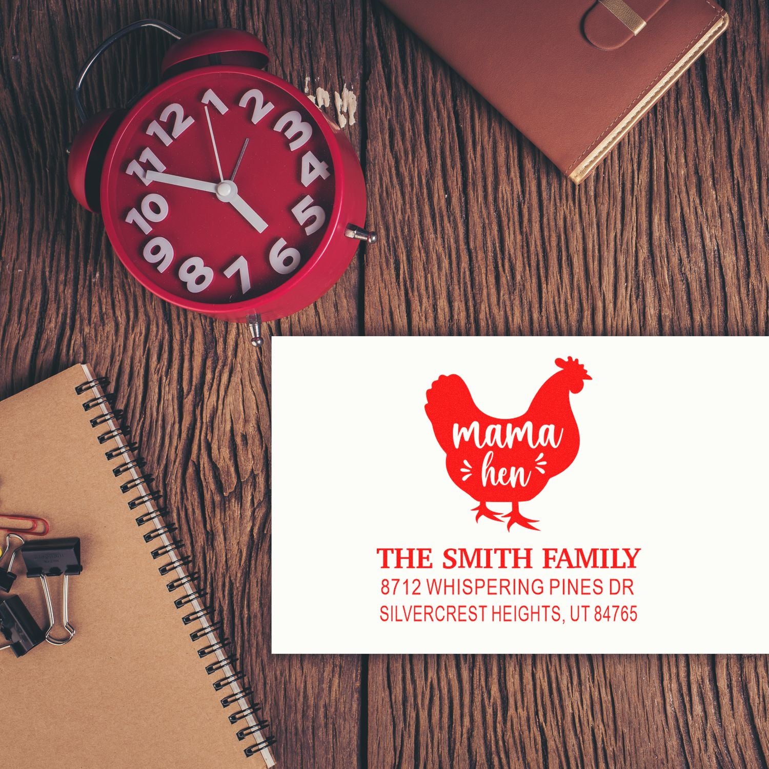 Custom return address stamp with red mama hen design on a white card, styled on a rustic wood desk with clock, notebook, and planner.