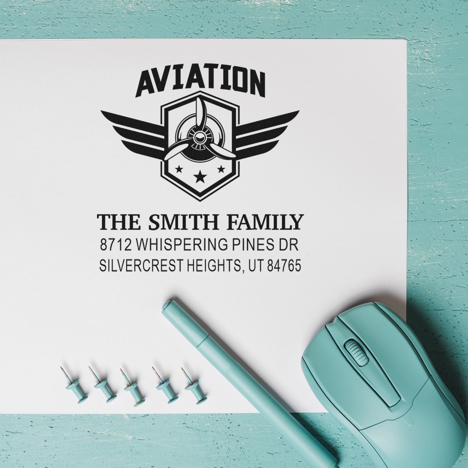 Custom aviation self-inking address stamp with winged propeller logo, The Smith Family Utah address, on white paper with teal stamp and pen.