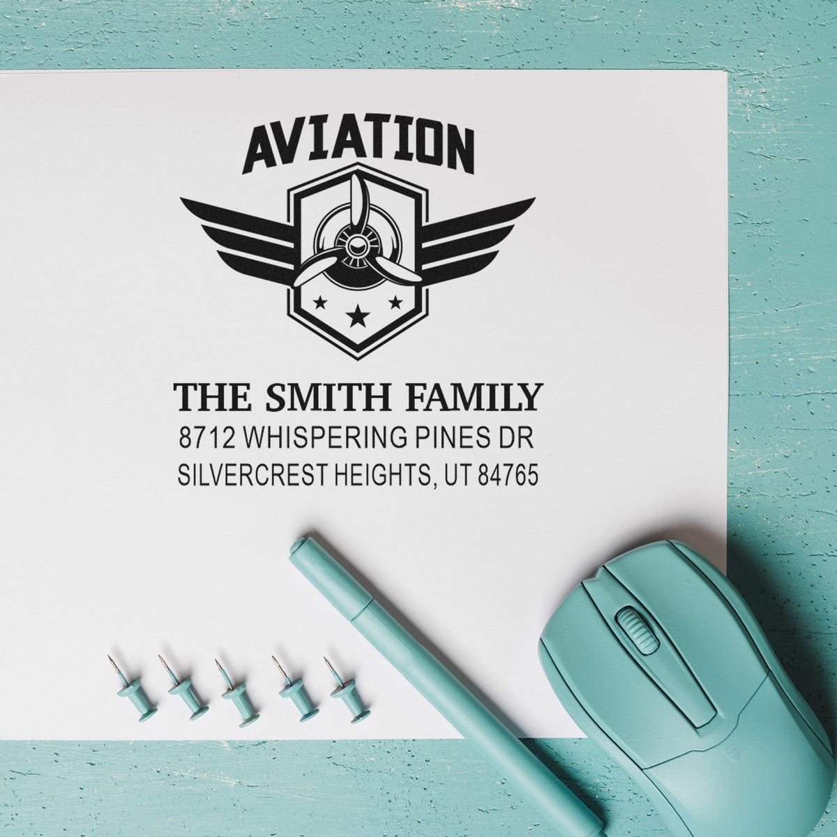Custom aviation family address stamp with airplane emblem on white paper, shown with teal stamp, pen, and replacement pins on a turquoise background.