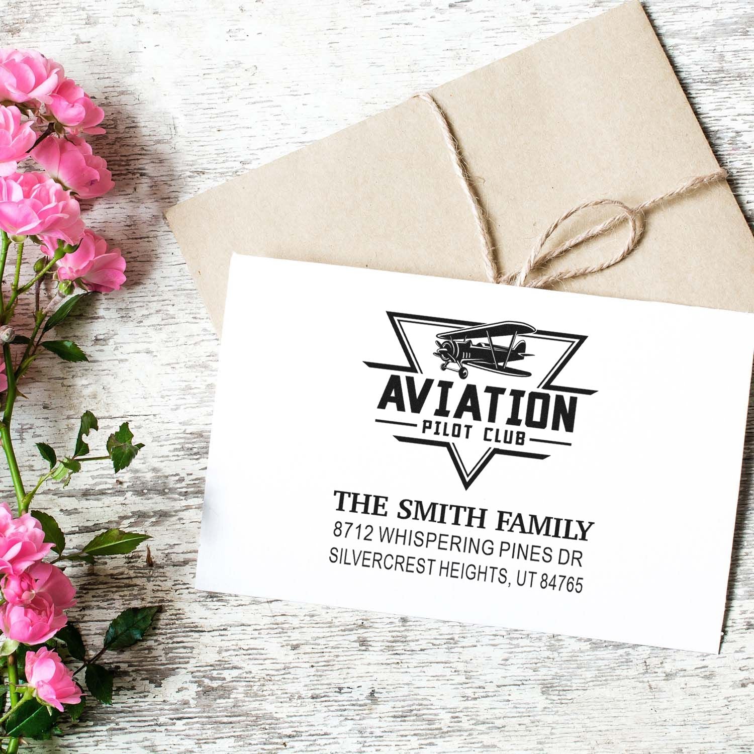 Custom aviation pilot club return address stamp on white paper beside a twine-wrapped kraft envelope and pink flowers on rustic wood table.