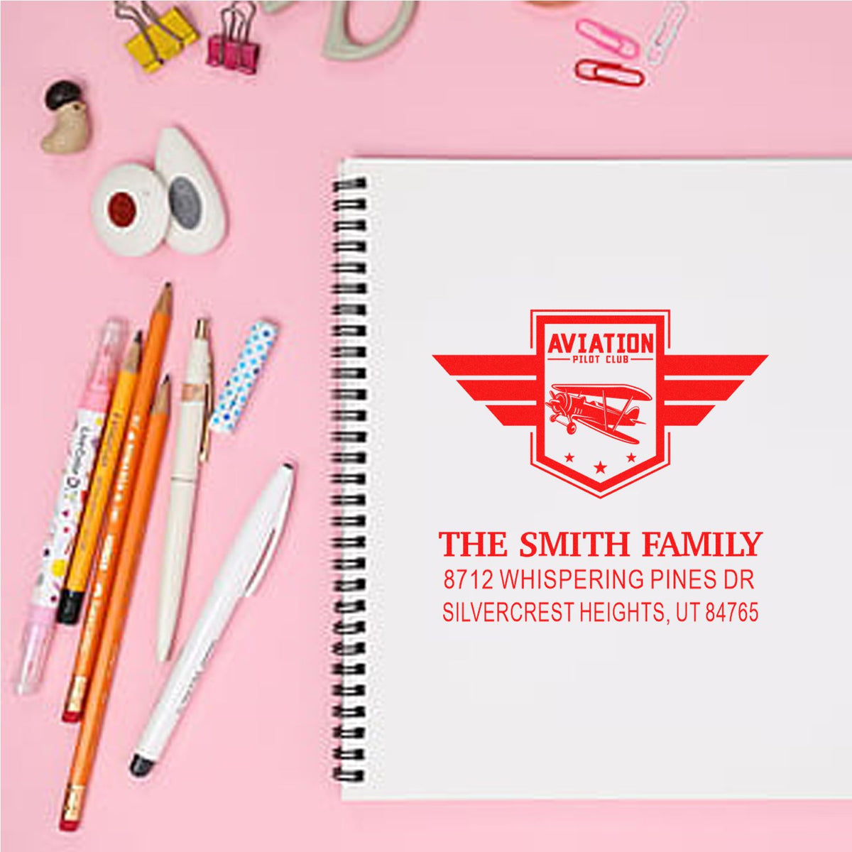 Custom aviation pilot club address stamp on a spiral notebook, styled on a pink desk with pens and office supplies.
