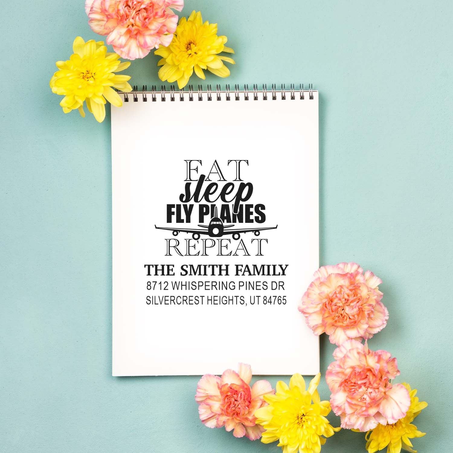 Custom family address stamp on a spiral notepad reading Eat Sleep Fly Planes Repeat, framed by pink and yellow flowers on a mint background.