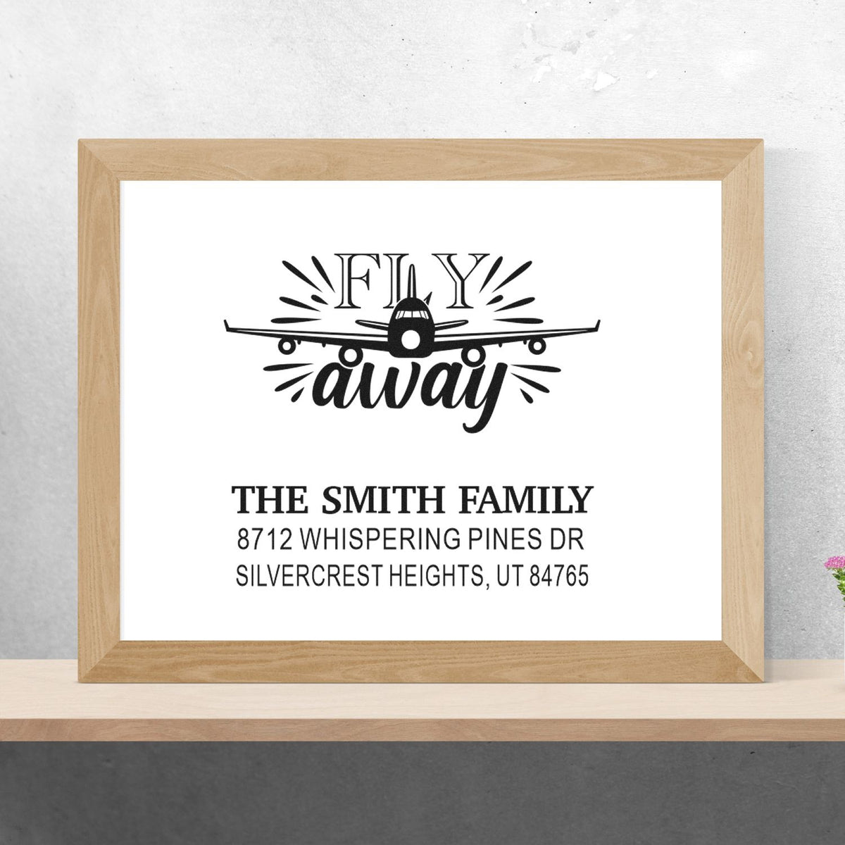Custom family address stamp with airplane and Fly away design, displayed in a framed mockup on a shelf.