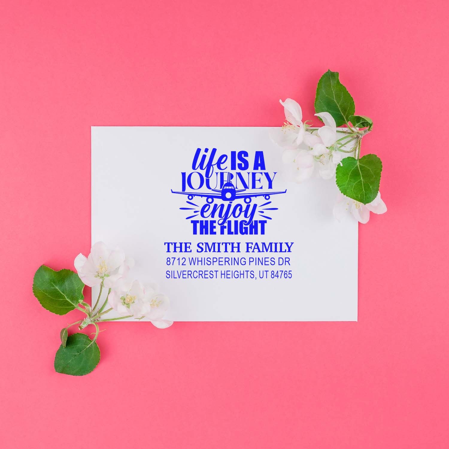 Custom address stamp with blue Life Is a Journey, Enjoy the Flight design on white card, styled with blossoms on a pink background.