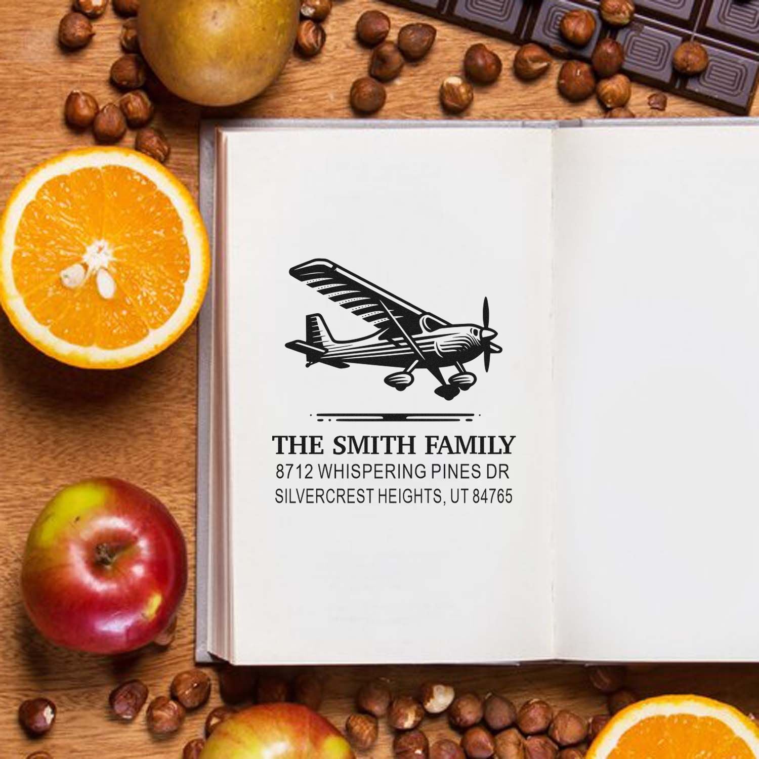 Custom family address stamp with vintage airplane on an open book, surrounded by oranges, apples, nuts, and a wooden tabletop.