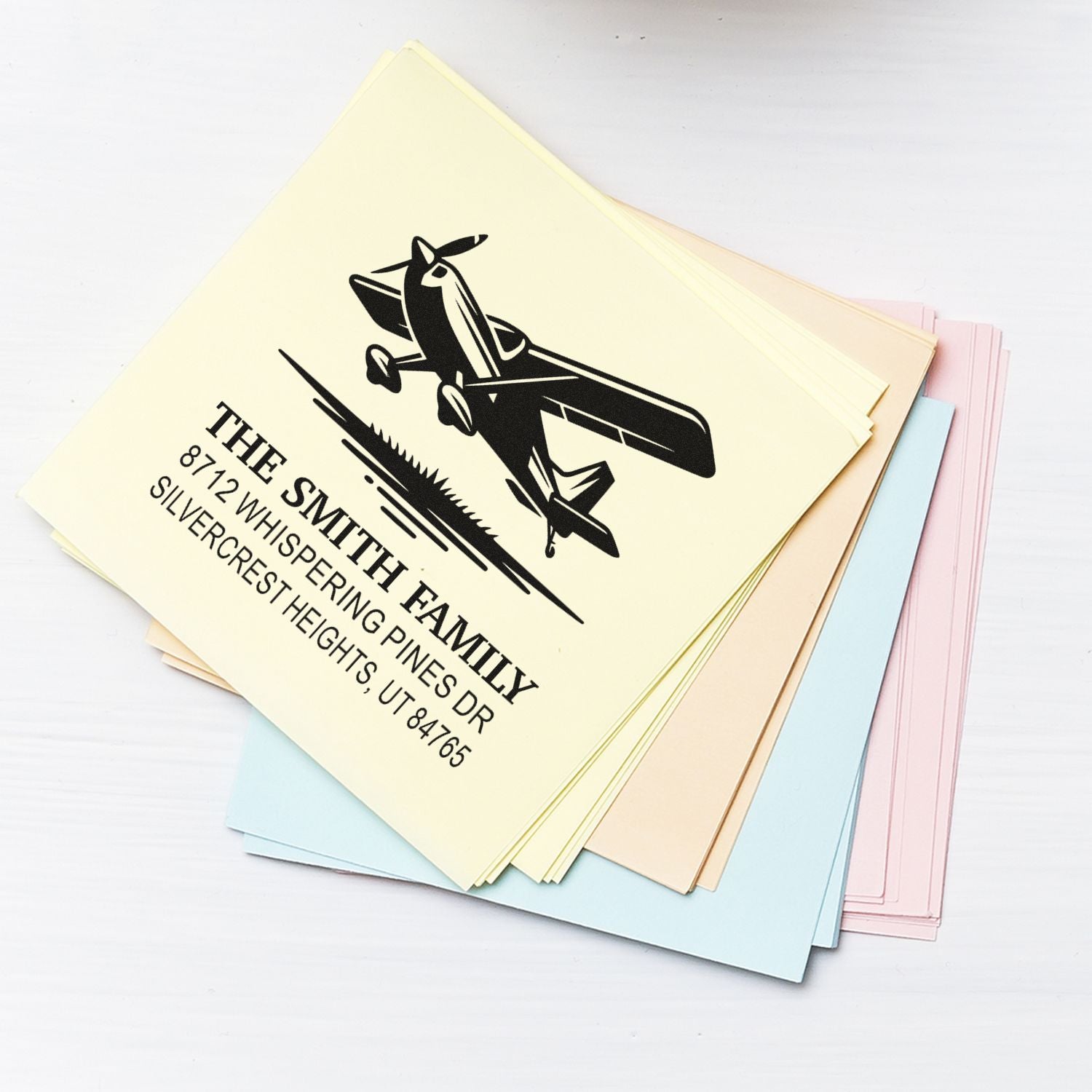 Custom self-inking stamps stacked on pastel pads, featuring an airplane logo and return address for The Smith Family on a white tabletop.