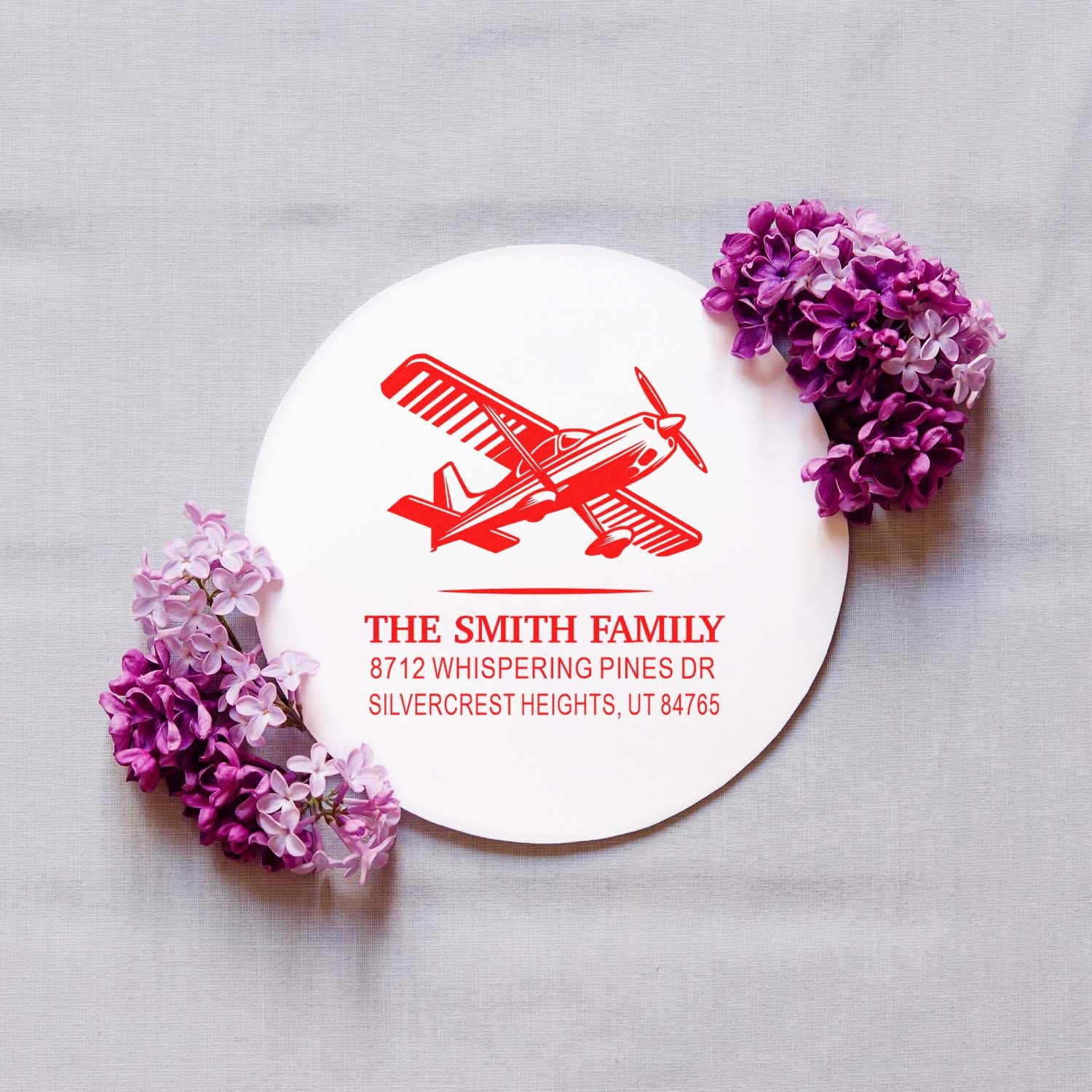 Custom red airplane family address stamp on round white card, accented with purple lilac flowers on a gray fabric background.