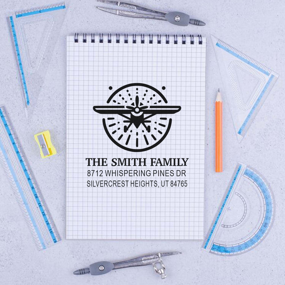 Custom family address stamp with airplane logo and home address on a notepad, surrounded by drafting tools, rulers, compasses, and a pencil on a desk.