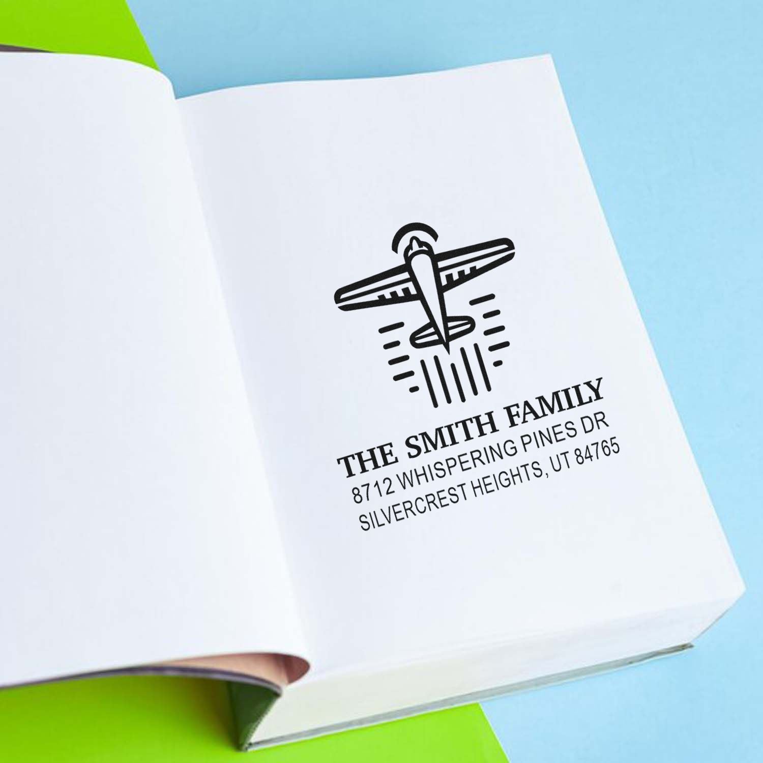 Personalized family address stamp with airplane design on open white paper, styled on a blue and green background.
