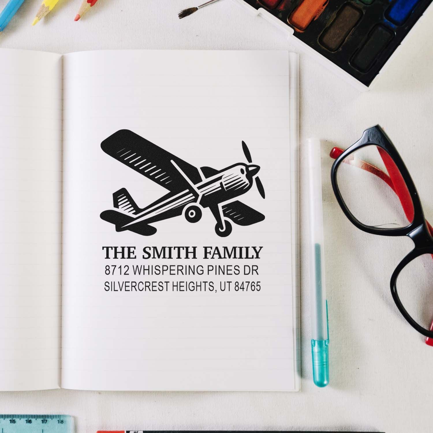 Custom airplane address stamp on an open notebook, showing The Smith Family Utah address, with art supplies, glasses and paint around it.