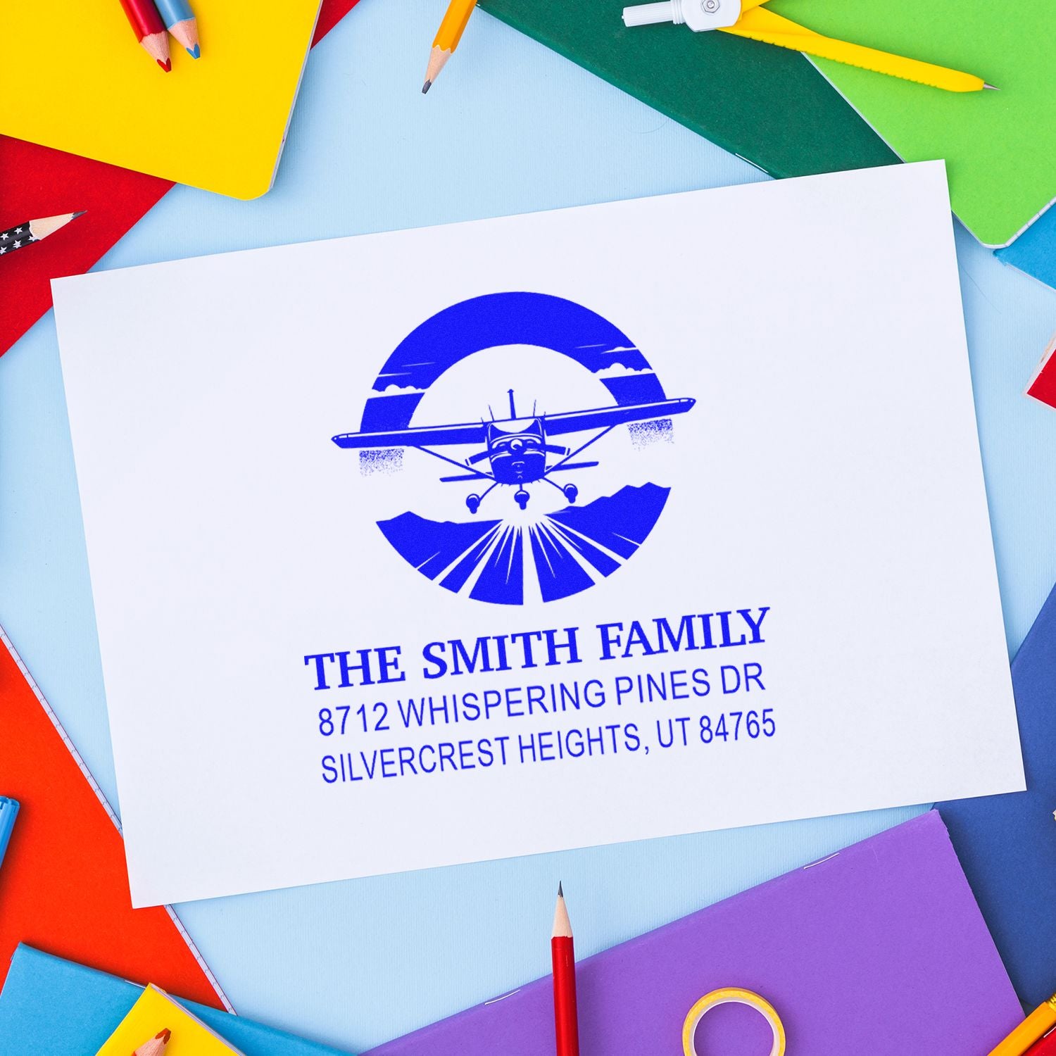 Personalized blue airplane return address stamp for The Smith Family on white card, surrounded by colorful notebooks and pencils.
