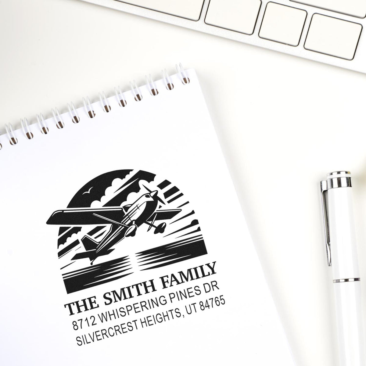 Personalized family address stamp with airplane design on a white notebook beside a pen and keyboard on a clean desk.