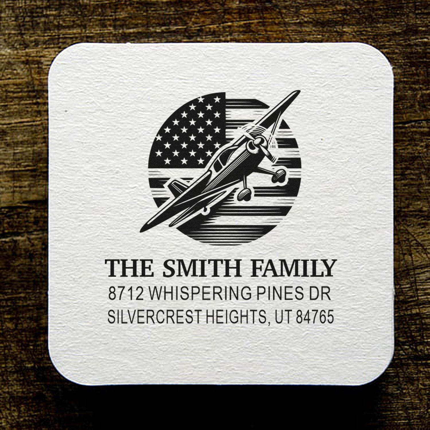 Personalized family address stamp with airplane and American flag design on white textured paper, shown on a rustic wood background.
