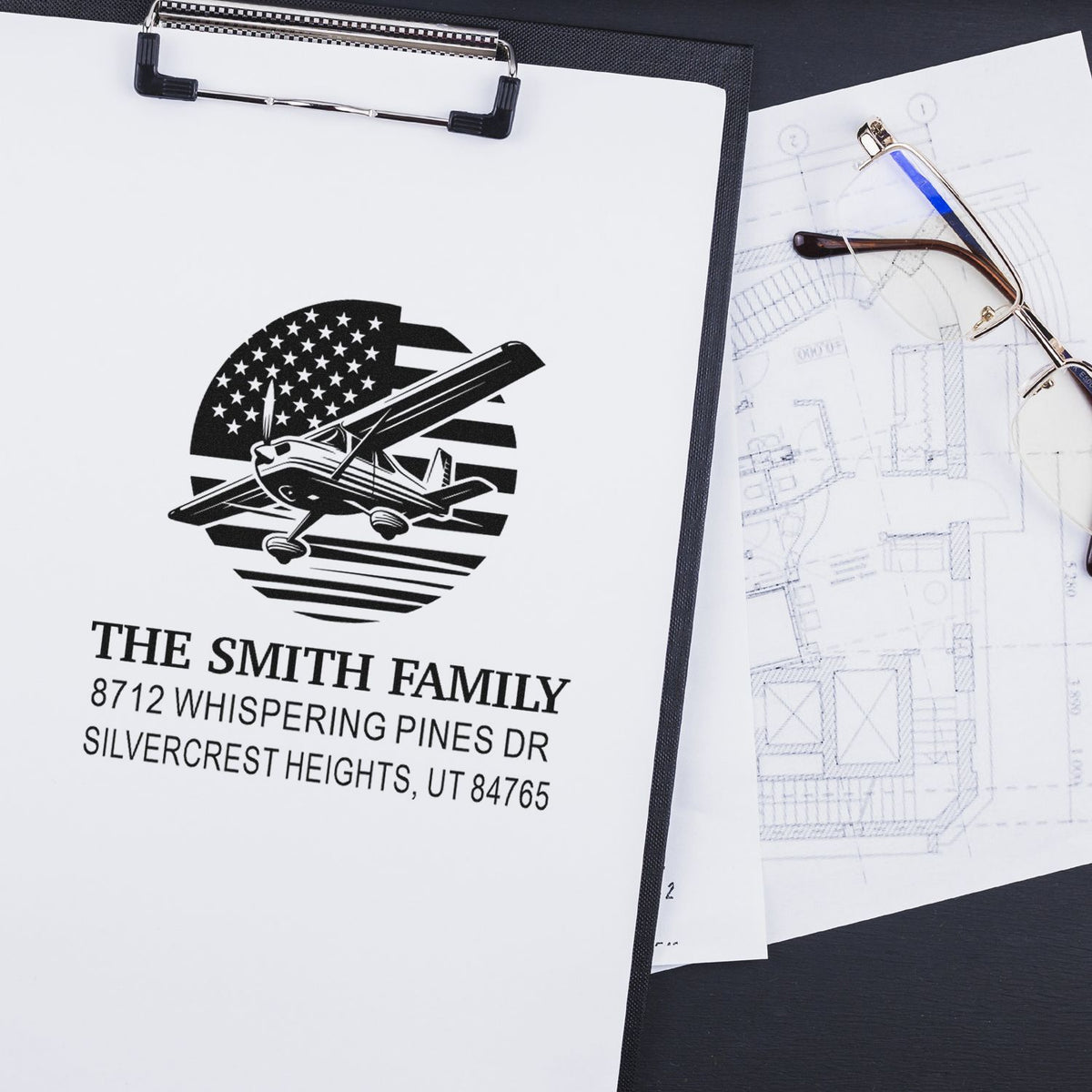 Custom address stamp with airplane logo and The Smith Family name on clipboard, beside eyeglasses and architectural plans.