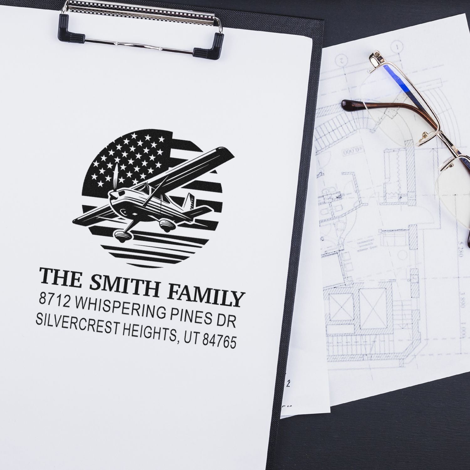 Custom rubber address stamp with airplane design and family address on a clipboard, beside eyeglasses and house blueprints on a desk.