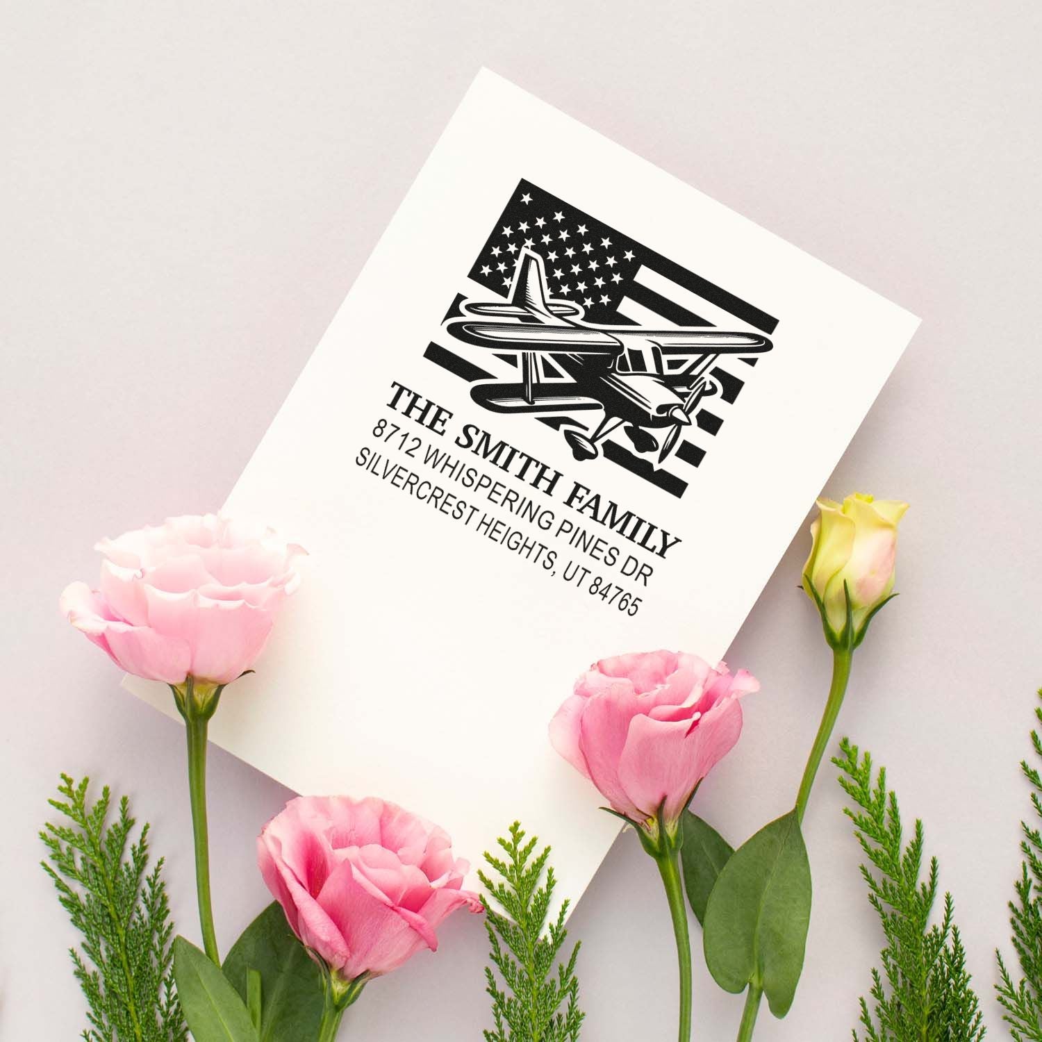 Custom family address stamp featuring airplane and US flag design on white card, surrounded by pink flowers on a soft pastel backdrop.