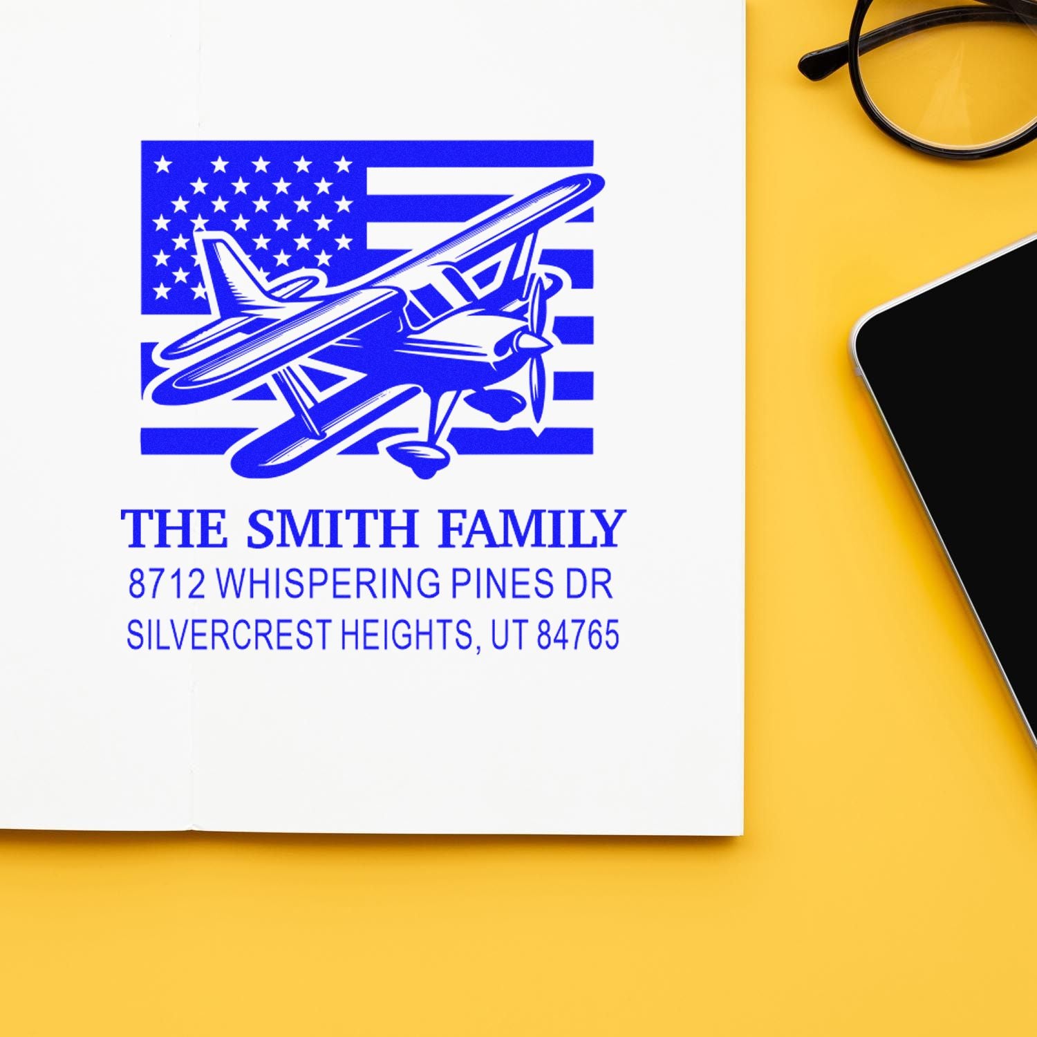Custom family address stamp with airplane and American flag design on white paper, beside glasses and smartphone on a yellow desk.