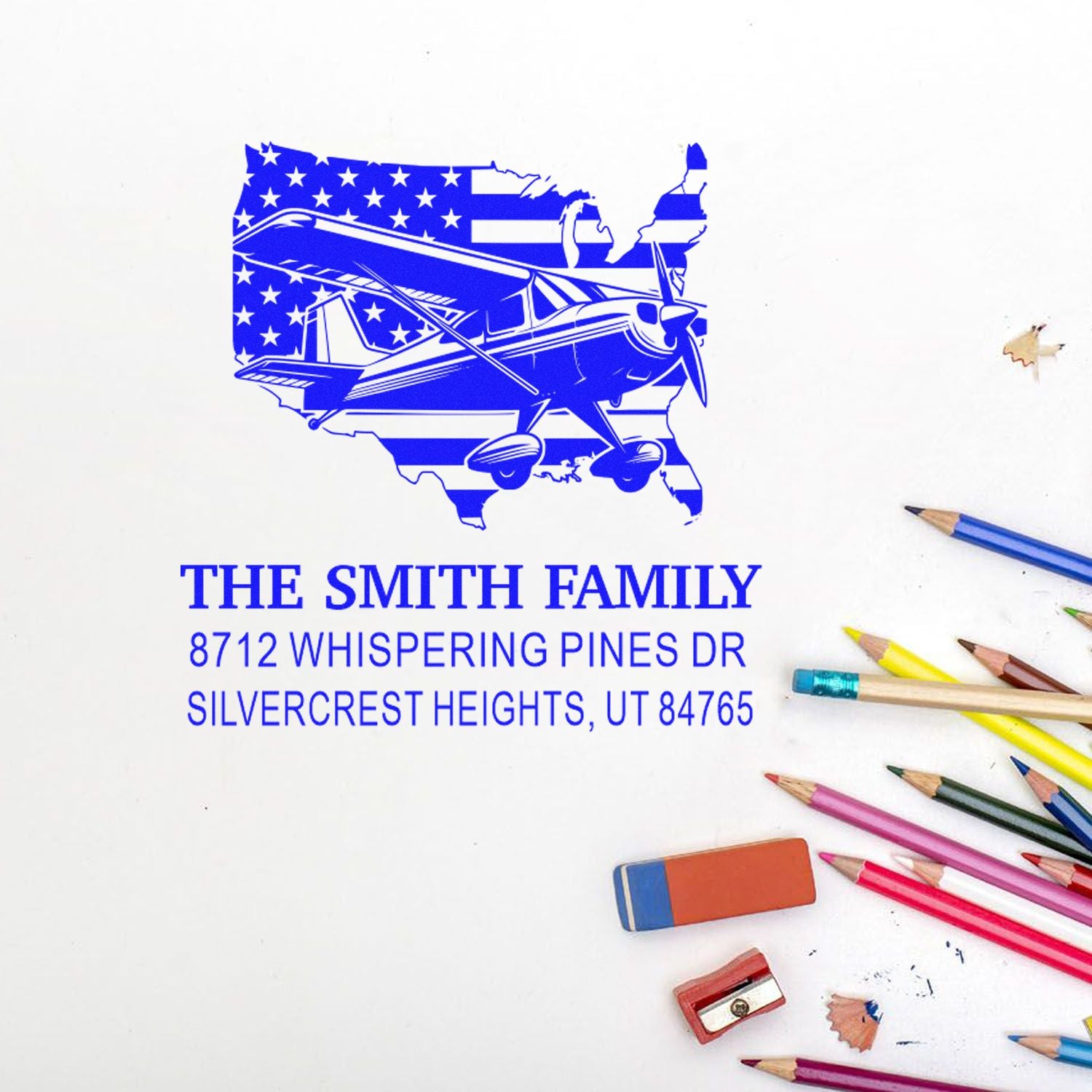 Personalized family address stamp with airplane over USA flag, featuring The Smith Family and Silvercrest Heights, UT, beside colored pencils.