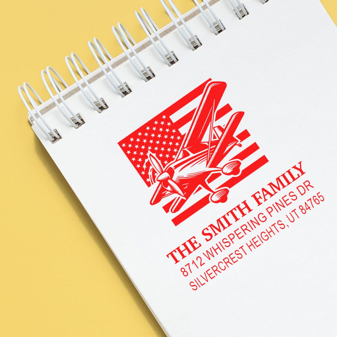 Custom family address stamp with airplane and U.S. flag design on a white spiral notebook against a yellow background.