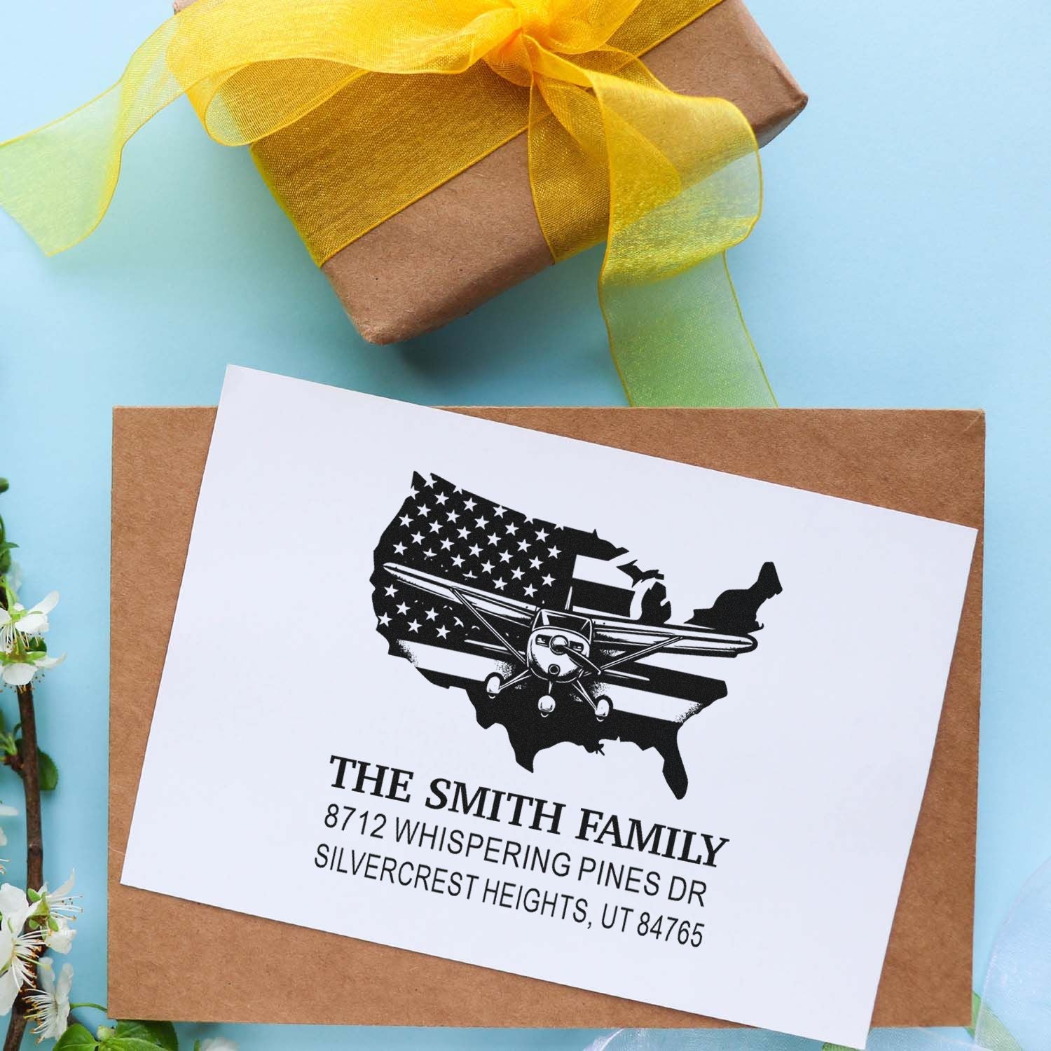 Custom return address stamp with U.S. map and airplane design on kraft envelope, beside a gift wrapped with yellow ribbon.