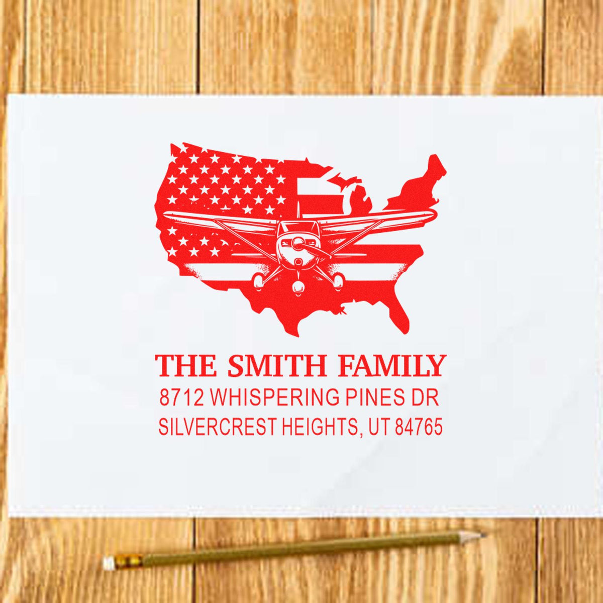 Custom red family address stamp with USA map and airplane design on white paper, resting on a wooden desk with pencil.