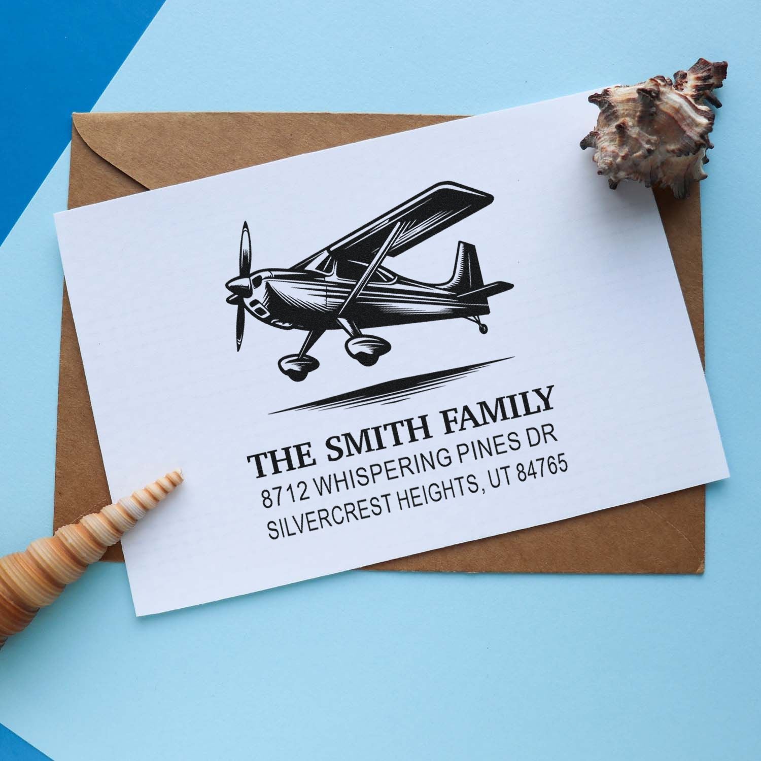 Personalized family address stamp with airplane design on white card over kraft envelope, styled with seashell accents on blue background.