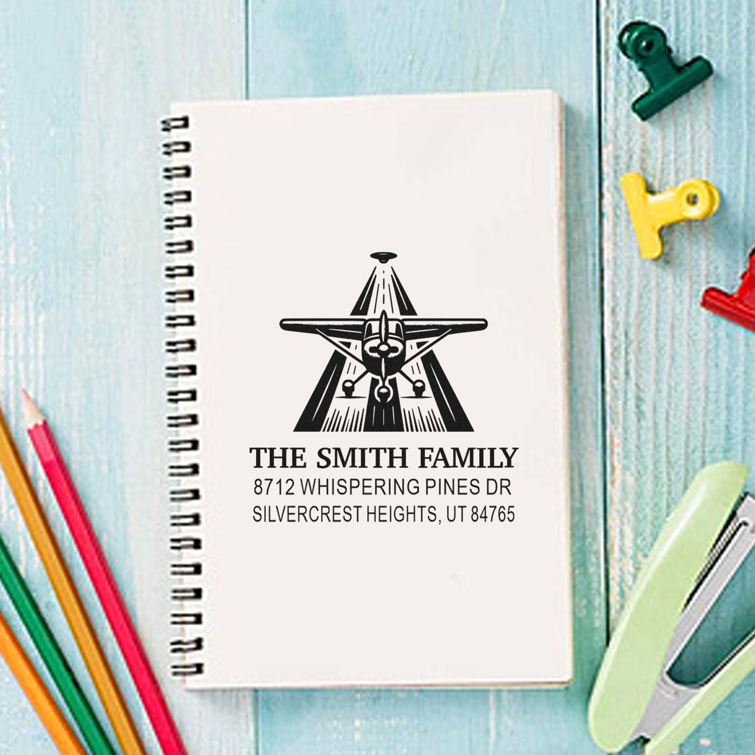 Personalized aviation address stamp on spiral notebook, with pencils, binder clips and colorful paper clips on a blue wood desk.