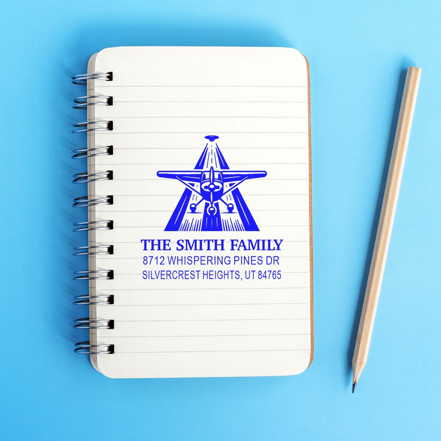 Custom blue family address stamp on a spiral notebook with pencil on a blue background, featuring an airplane logo and Smith family address.
