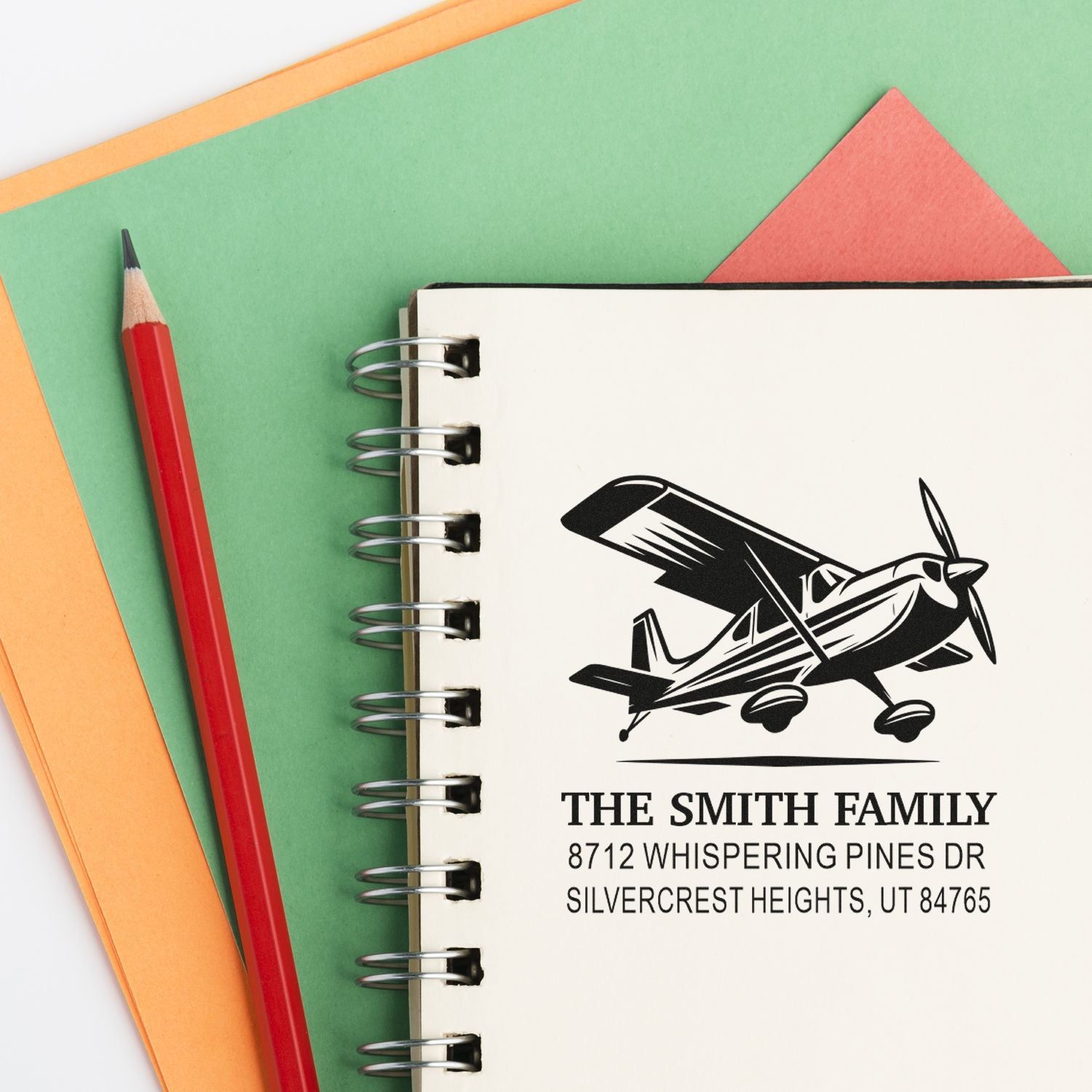 Personalized family address stamp with airplane design on spiral notebook, beside a red pencil and colorful paper background.