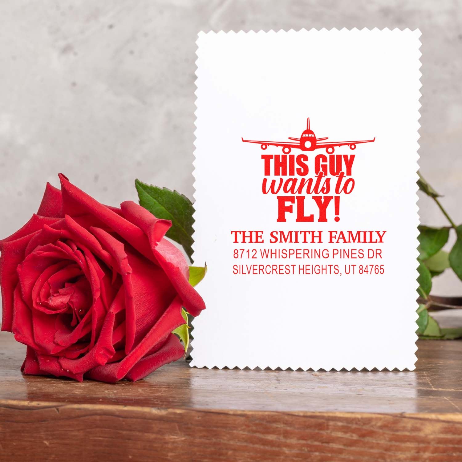 Personalized aviation-themed rubber stamp reading This Guy Wants to Fly! beside a red rose on a wooden table.