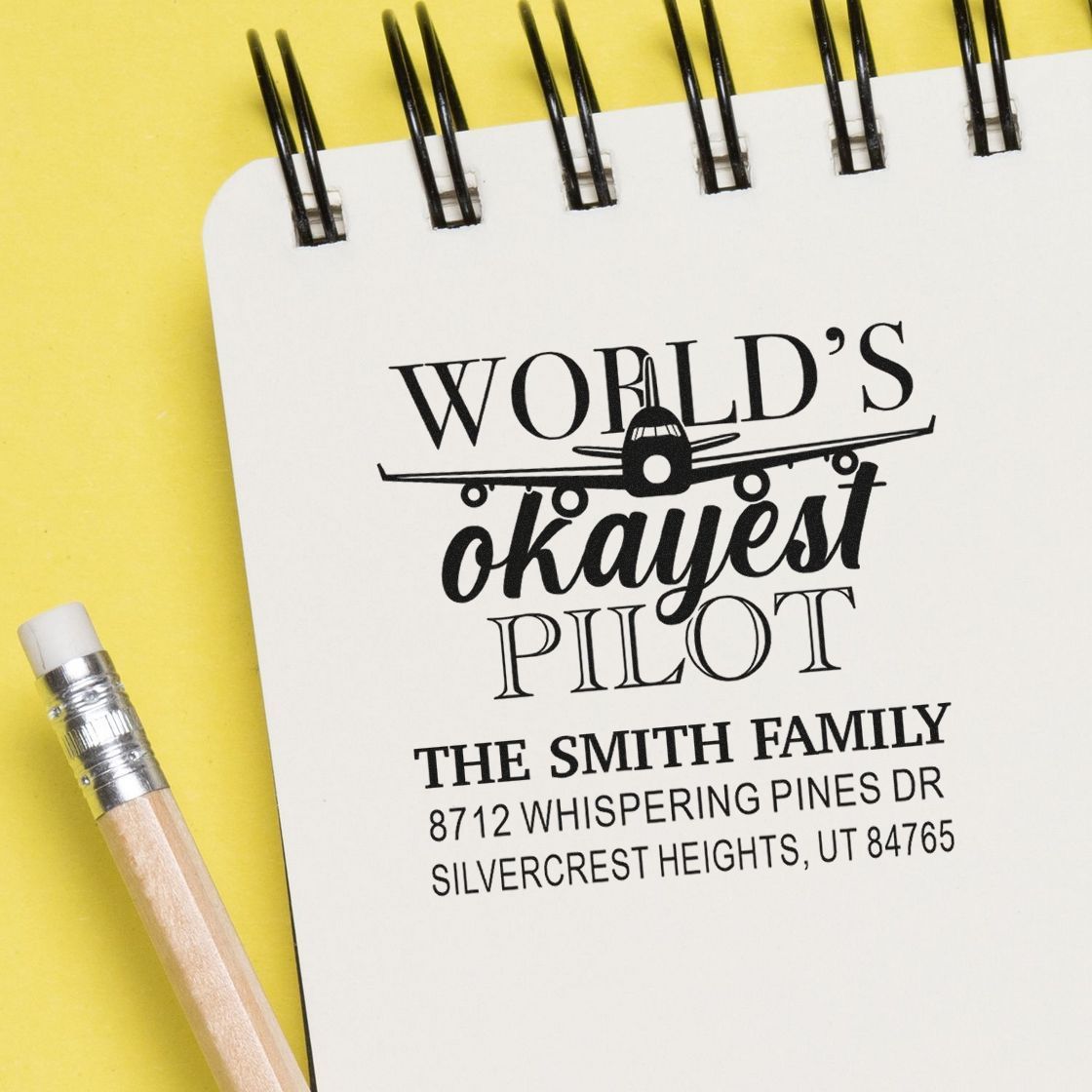 Custom rubber address stamp with Worlds Okayest Pilot airplane design, featuring family name and return address on a notebook page.