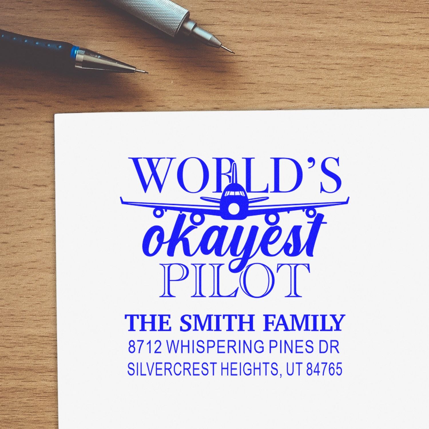 Custom pilot address stamp on white paper, reading Worlds Okayest Pilot, with airplane graphic, on a wooden desk beside two pens.