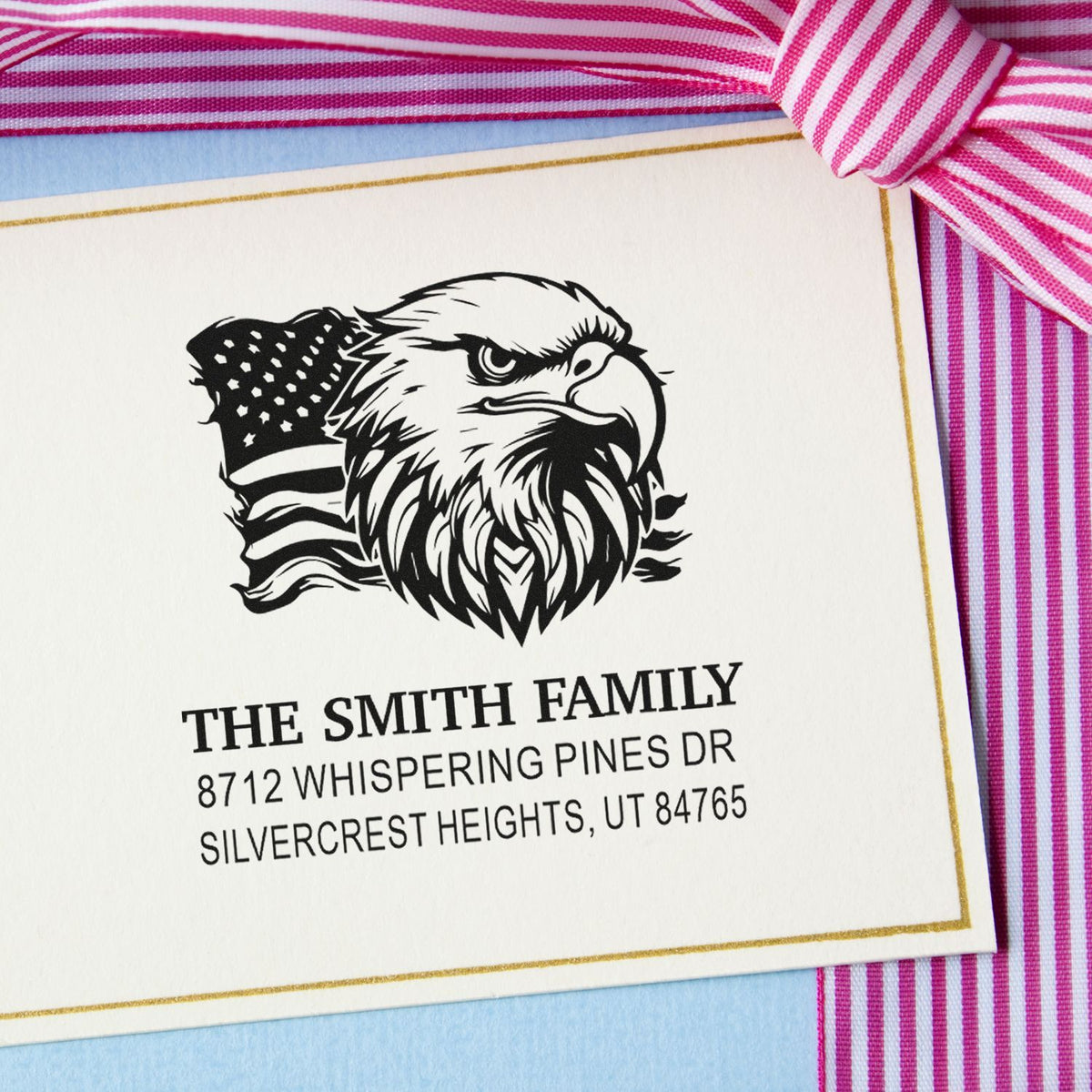 Custom return address stamp with eagle and American flag design on white card, styled with pink striped ribbon and blue background.