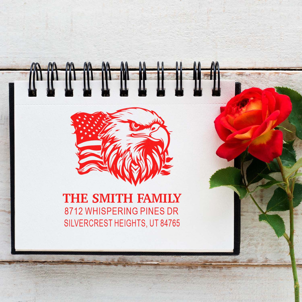 Custom family address stamp with eagle and American flag design on a spiral notepad beside a red rose on rustic wood.