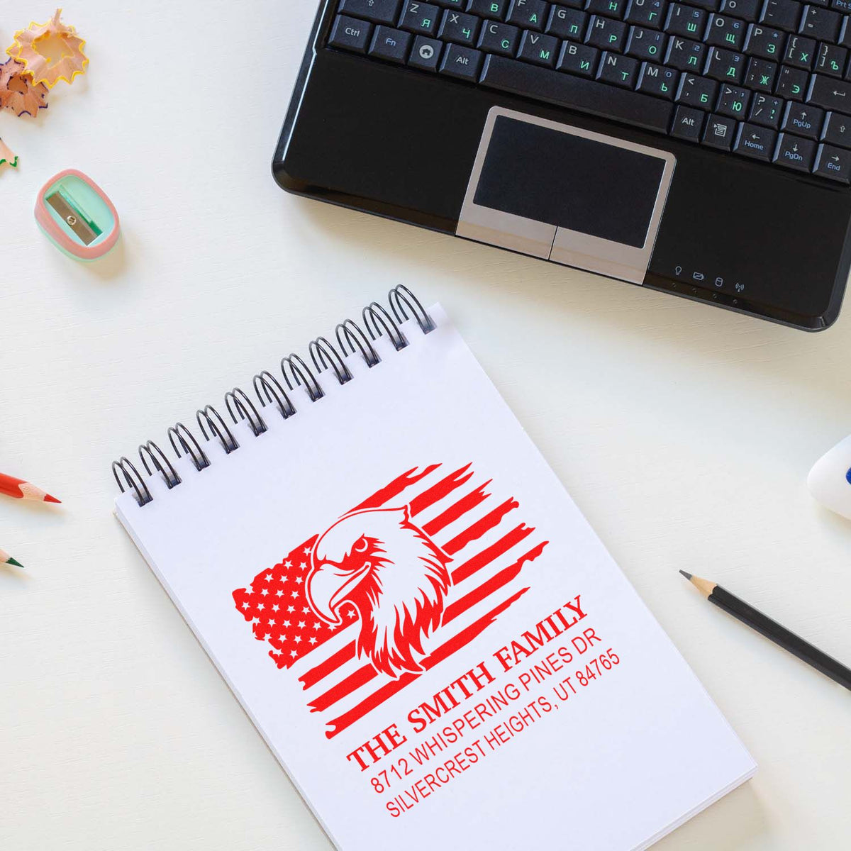 Personalized family address stamp with eagle and American flag design on a spiral notebook beside a laptop on a white desk.
