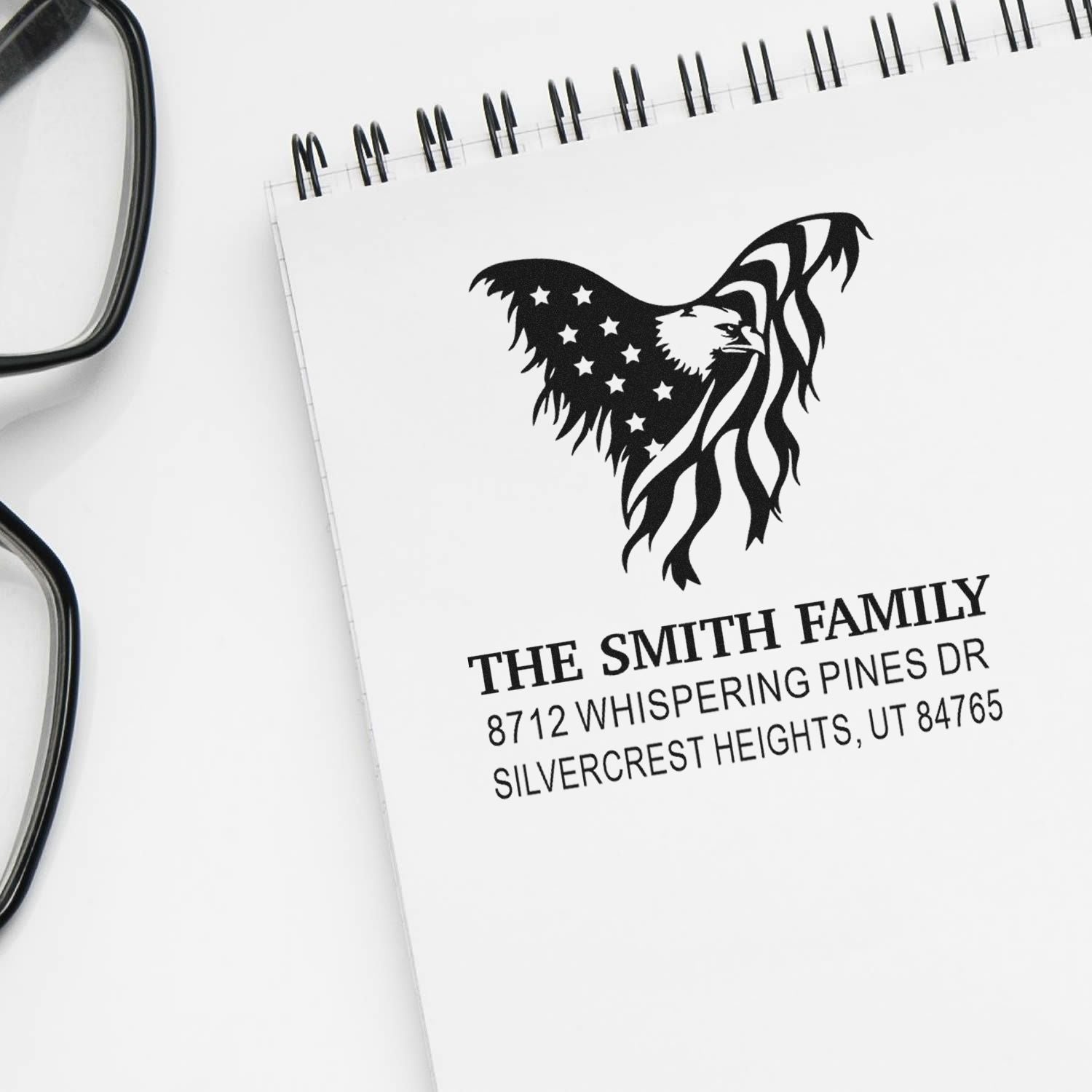 Custom family address stamp on a spiral notebook, featuring patriotic eagle and American flag design beside eyeglasses.