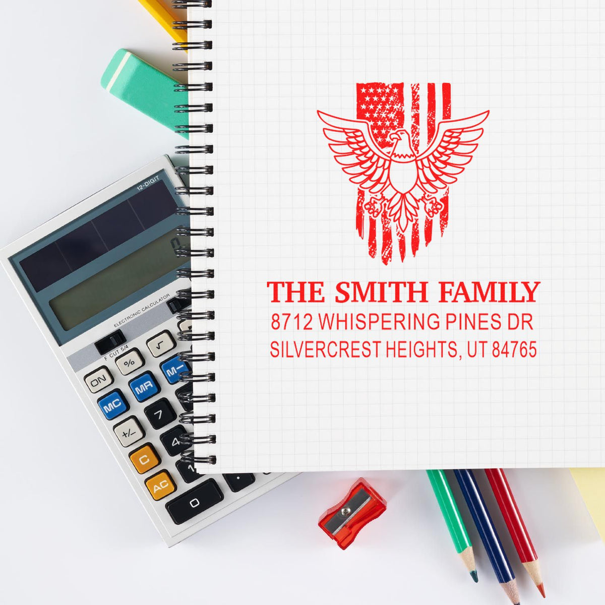 Custom family address rubber stamp with eagle and American flag design, displayed on a notebook with calculator, pencils, and sharpener for office use.