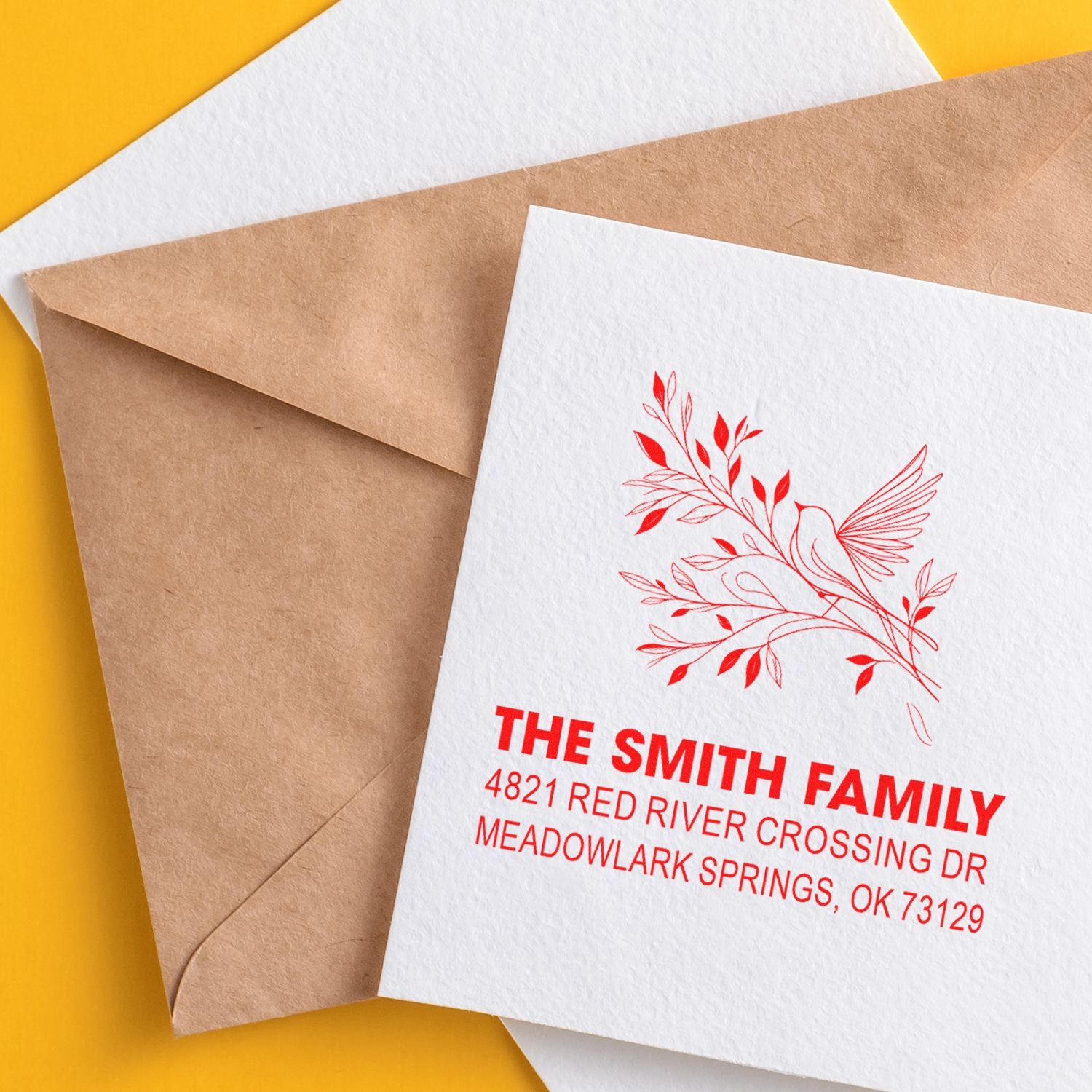 Custom return address rubber stamp with floral bird design, red ink on white envelope, shown with kraft mailer and blank stationery on yellow background.