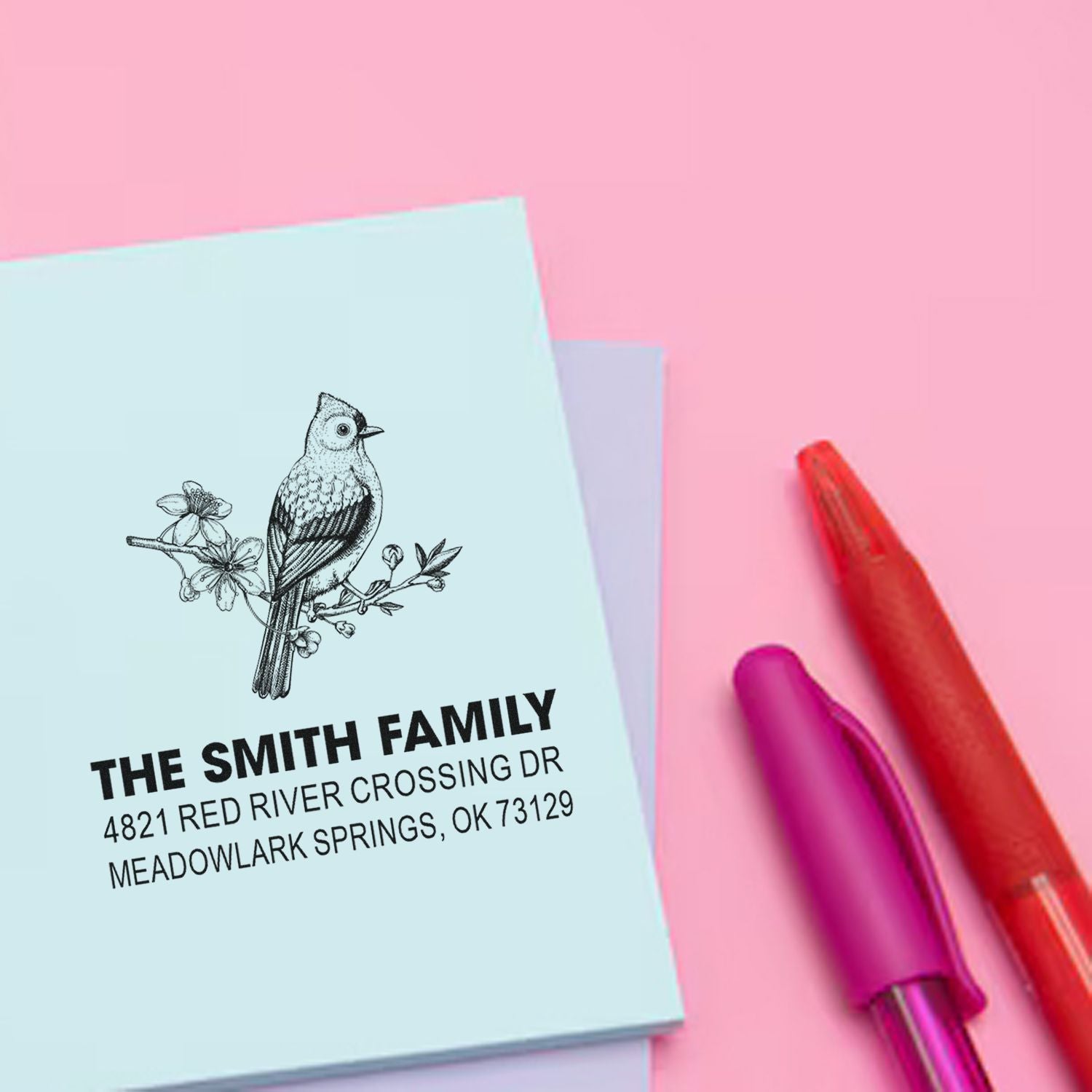 Custom rubber address stamp with bird and floral motif, featuring The Smith Family return address on pastel stationery beside colorful pens.