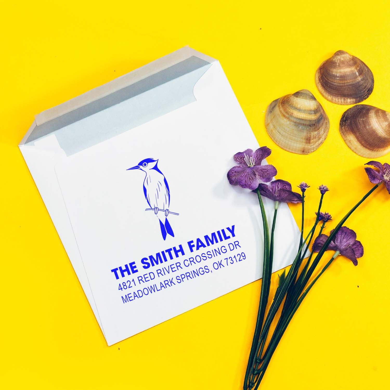 Custom return address stamp with blue bird design and The Smith Family address on a white mailer box, styled with shells and purple flowers.