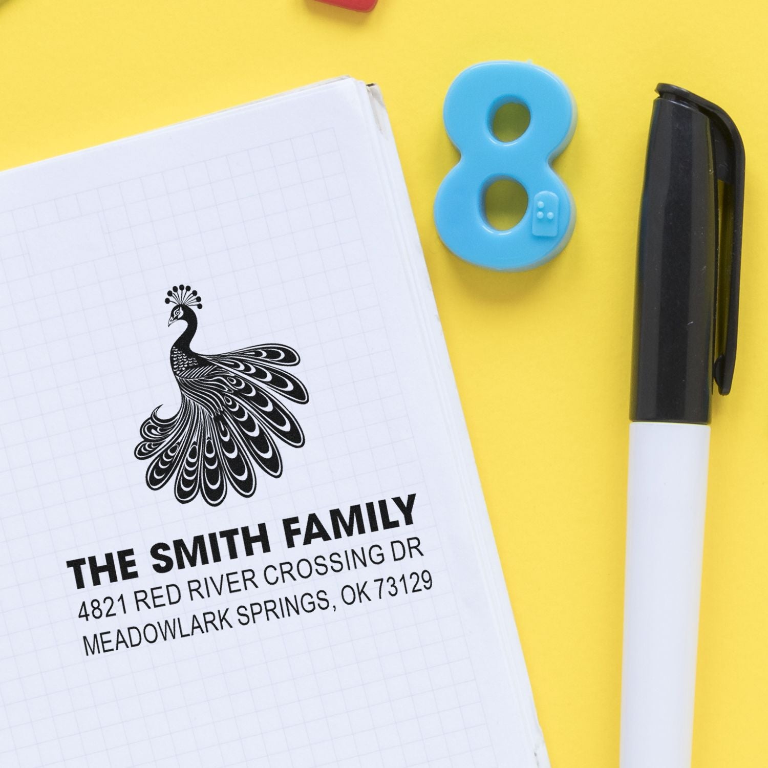 Custom self-inking address stamp with peacock design, personalized family name and return address, shown beside a black marker on a yellow background.