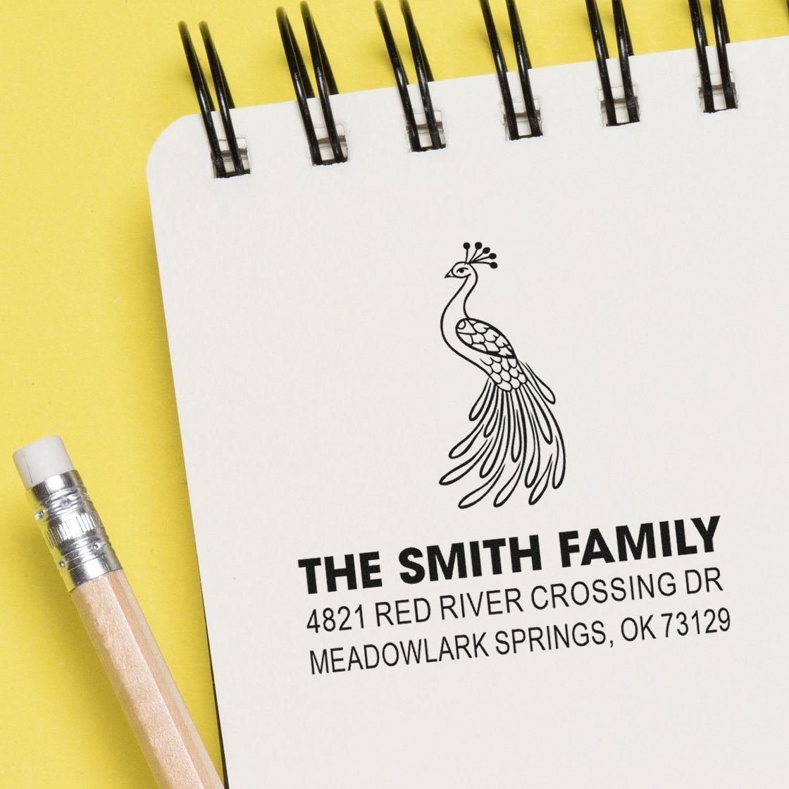 Custom rubber address stamp with peacock design, family name, and mailing address printed on a white spiral notebook beside a pencil on yellow background.