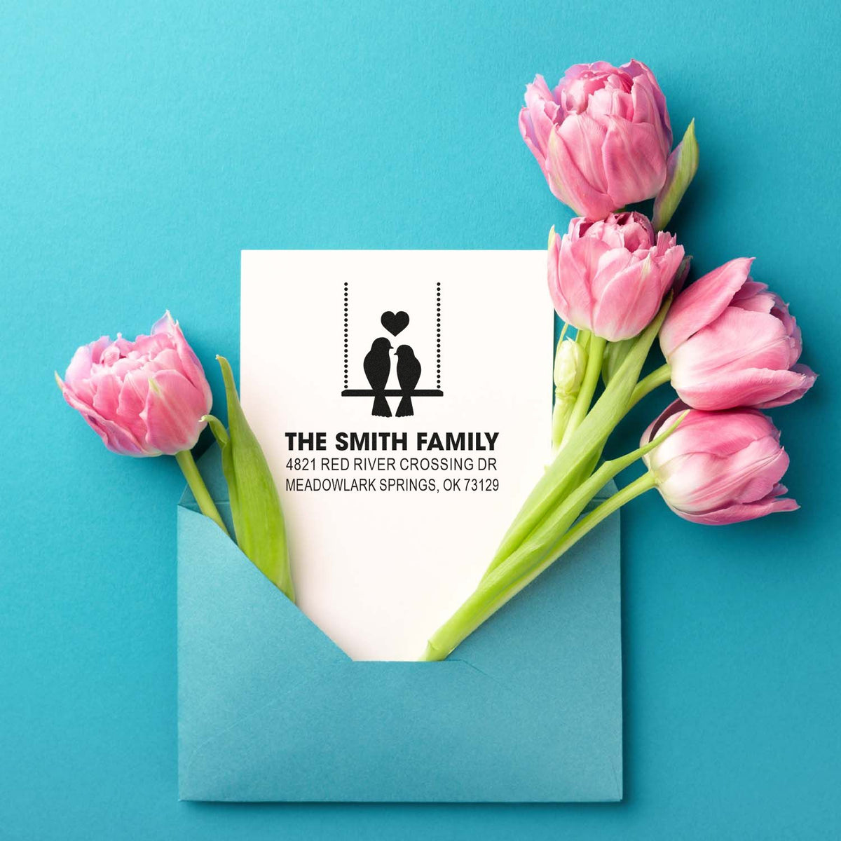 Personalized family return address rubber stamp with bird and heart motif, printed on envelope with pink tulips and blue background.