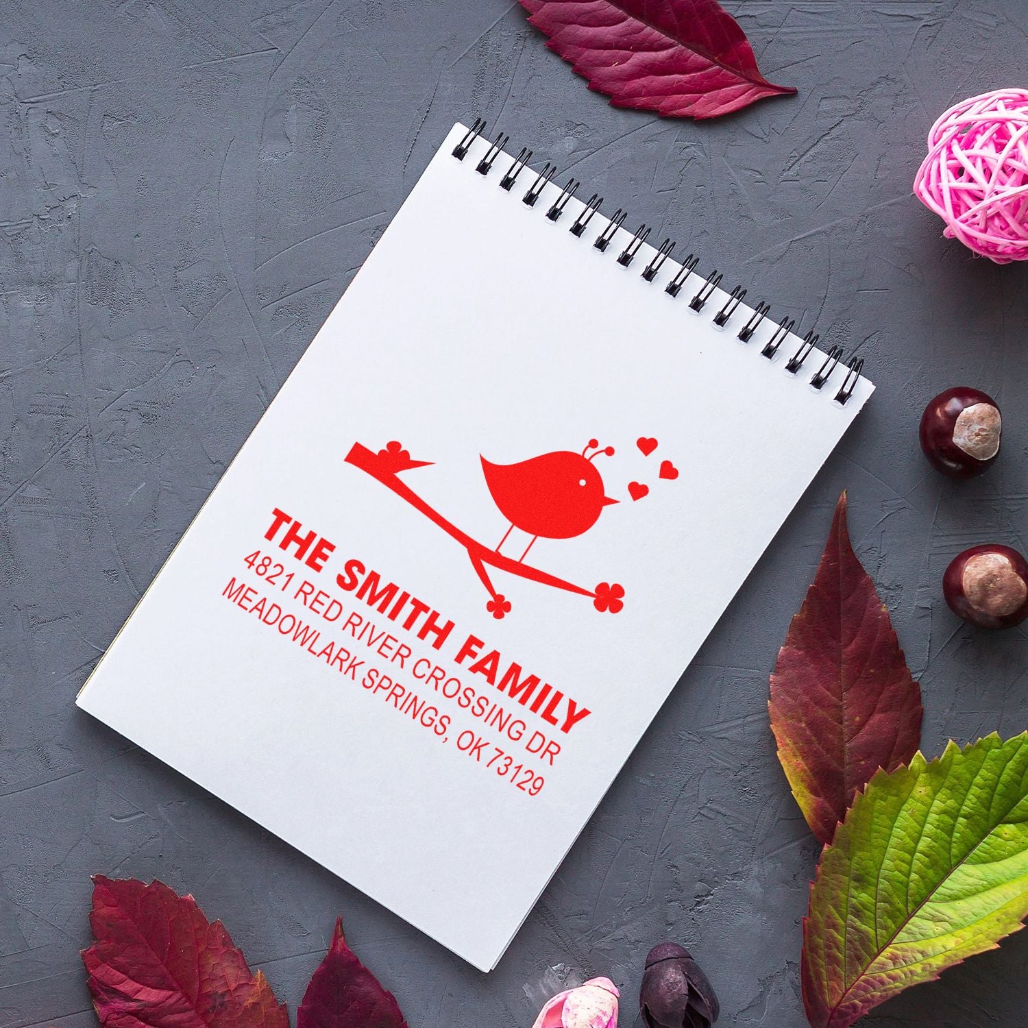 Custom return address rubber stamp with bird logo, personalized family name and address, red ink impression on spiral notebook paper