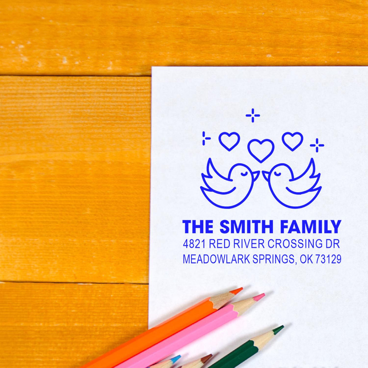 Personalized family return address rubber stamp with birds and hearts, blue imprint on white paper beside colored pencils on a wooden desk for mailings.