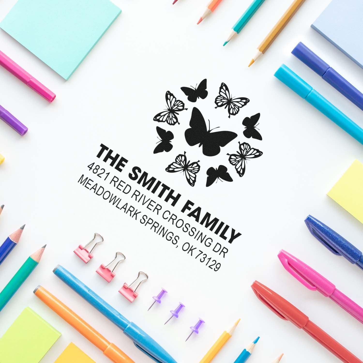 Personalized butterfly return address rubber stamp with custom family name and mailing address, surrounded by colorful pens and stationery supplies.